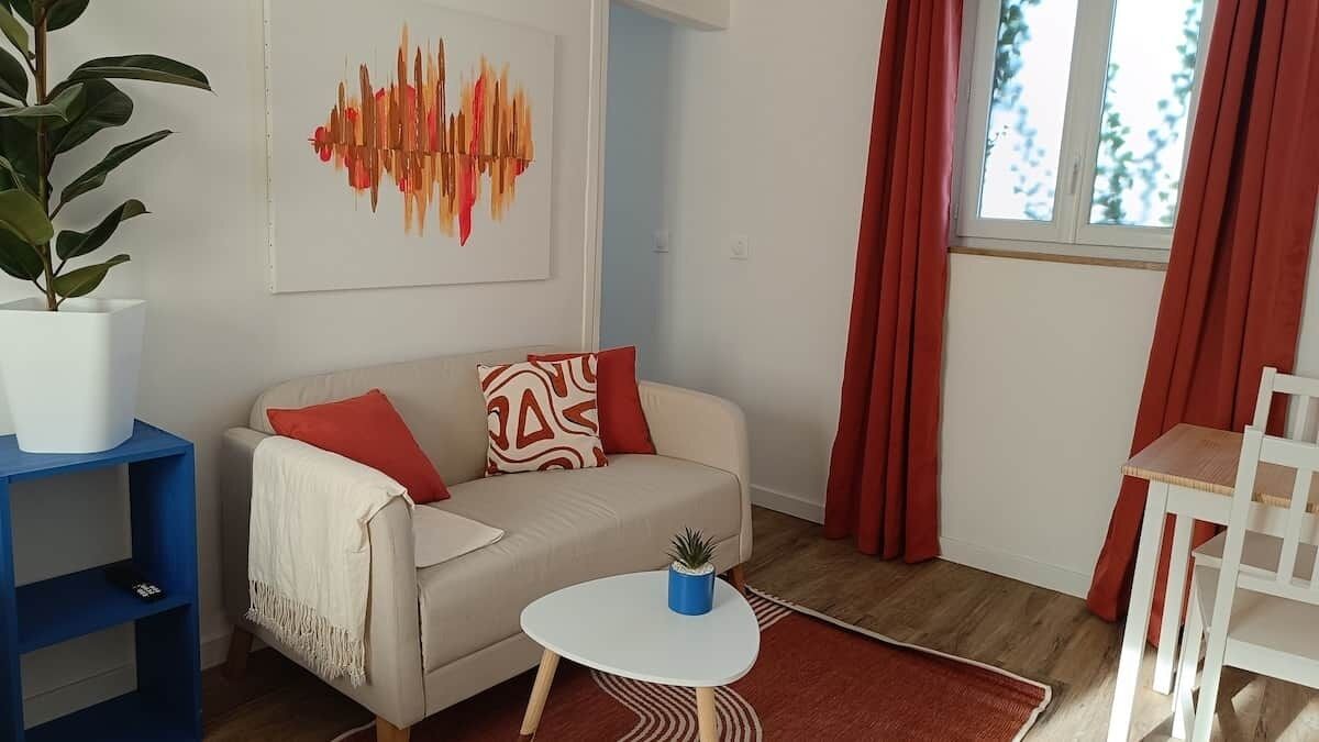 Un salon avec un canapé, une table basse, une plante, un tableau et une fenêtre avec des rideaux.