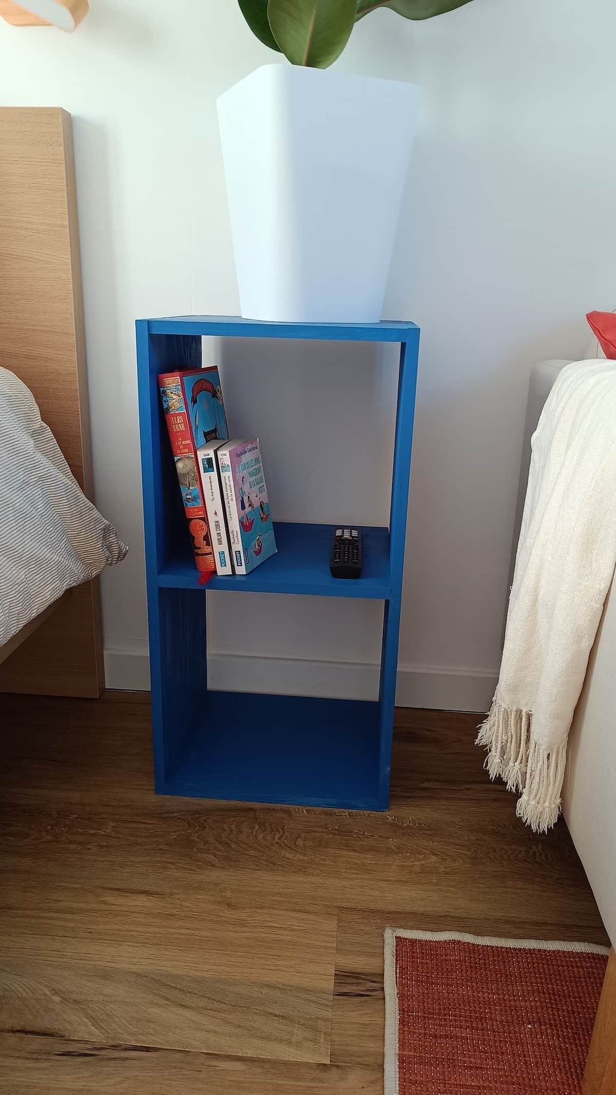 Een blauwe boekenkast met boeken en een afstandsbediening, een witte plant en een witte plaid op een houten vloer.