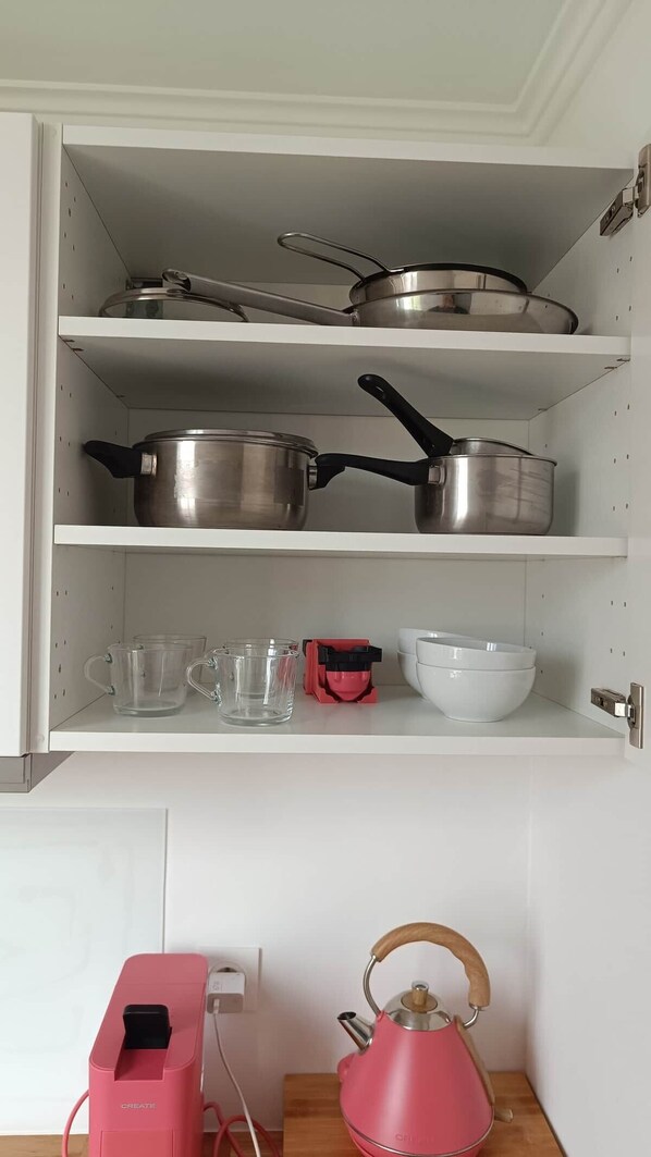 A kitchen with stainless steel cookware on shelves, clear drinking glasses, a red electric kettle, and a white mixing bowl.