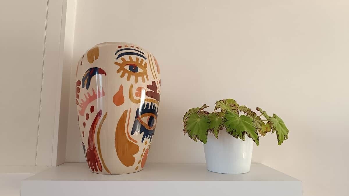 Un jarrón de cerámica con diseño facial y una planta en maceta sobre una repisa.