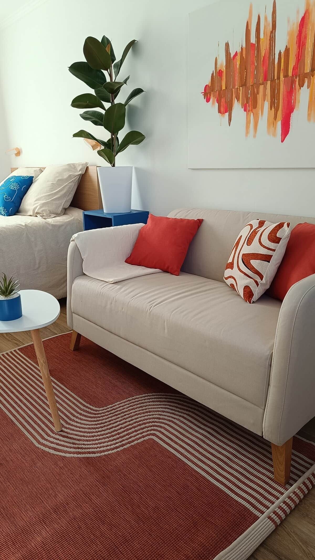 Een moderne woonkamer met een beige bank, rode en gemêleerde kussens, een grote plant en een kunstwerk aan de muur.