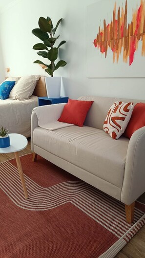 A modern living room with a beige sofa, red and patterned cushions, a large plant, and a wall art piece. - defaultName (Songeons)