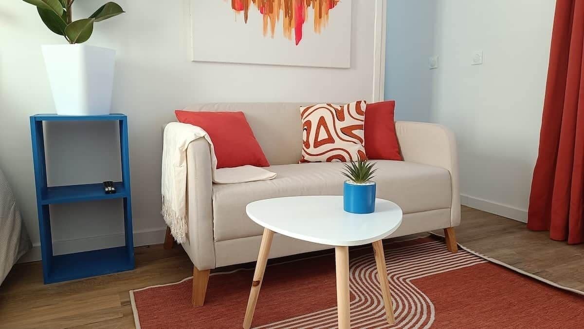 A living room with a sofa, a small table, a blue side table, a potted plant, a red curtain, and a patterned rug.