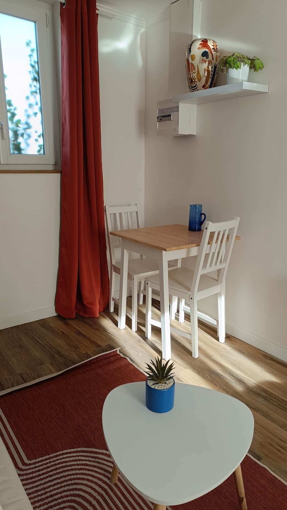 A dining area with a wooden table and white chairs, a red curtain, a potted plant, and a wall-mounted shelf with decorative items.