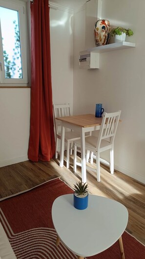 A dining area with a wooden table and white chairs, a red curtain, a potted plant, and a wall-mounted shelf with decorative items. - defaultName (Songeons)