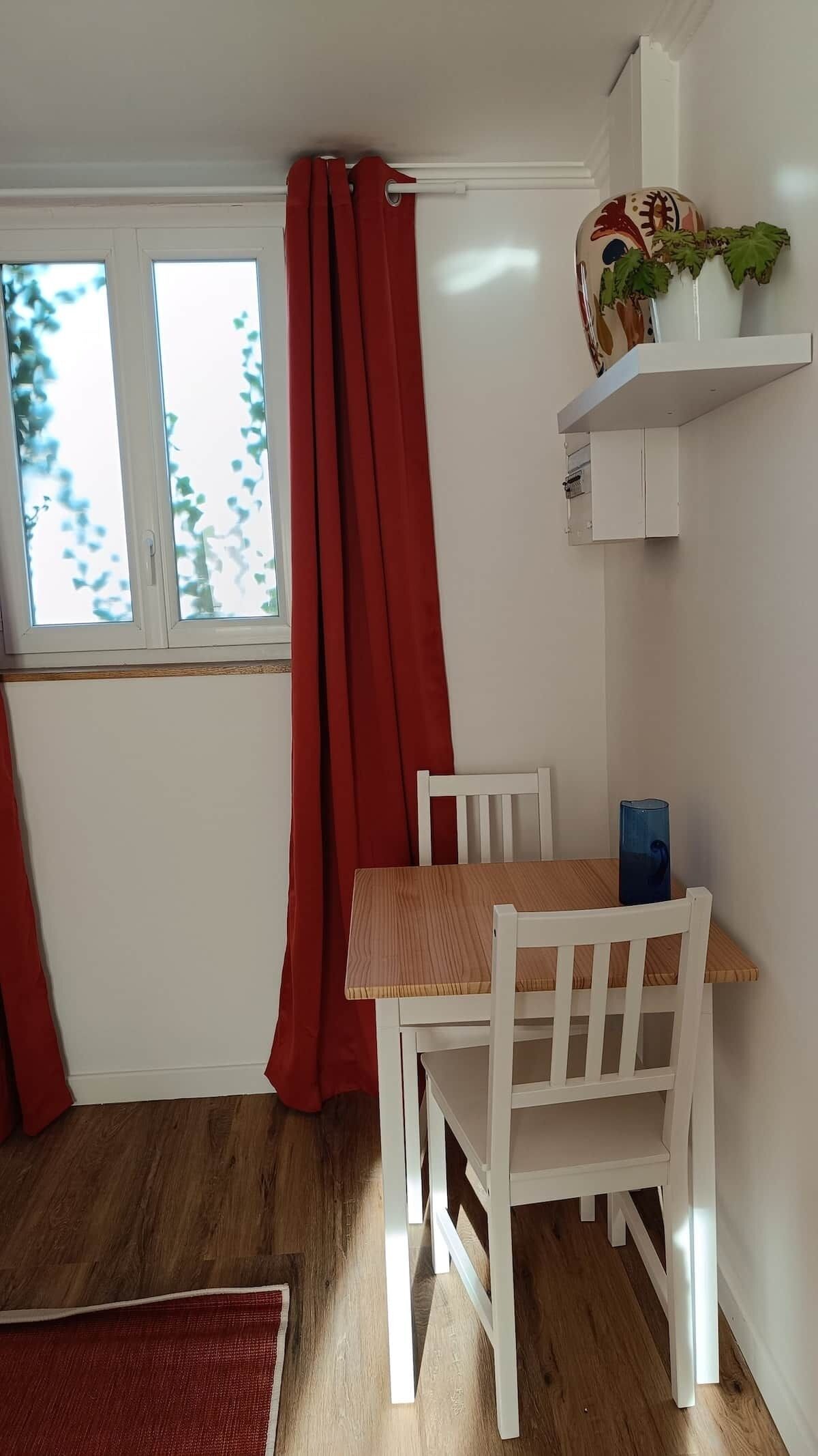 Een kamer met een houten tafel en witte stoelen, een raam met rode gordijnen en een plank met decoratieve objecten.