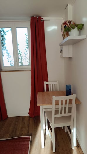 A room with a wooden table and white chairs, a window with red curtains, and a shelf with decorative items. - defaultName (Songeons)