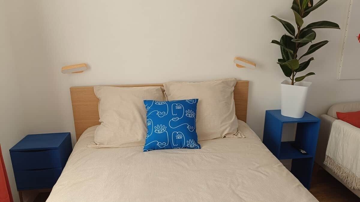 Een bed met hoofdbord, twee kussens, een nachtkastje en een potplant.