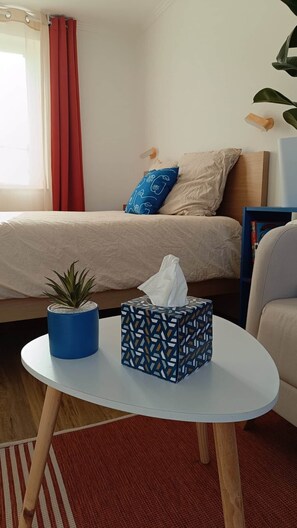 A bedroom with a bed, a bedside table with a tissue box and a plant, a window with curtains, and a wall-mounted lamp. - defaultName (Songeons)