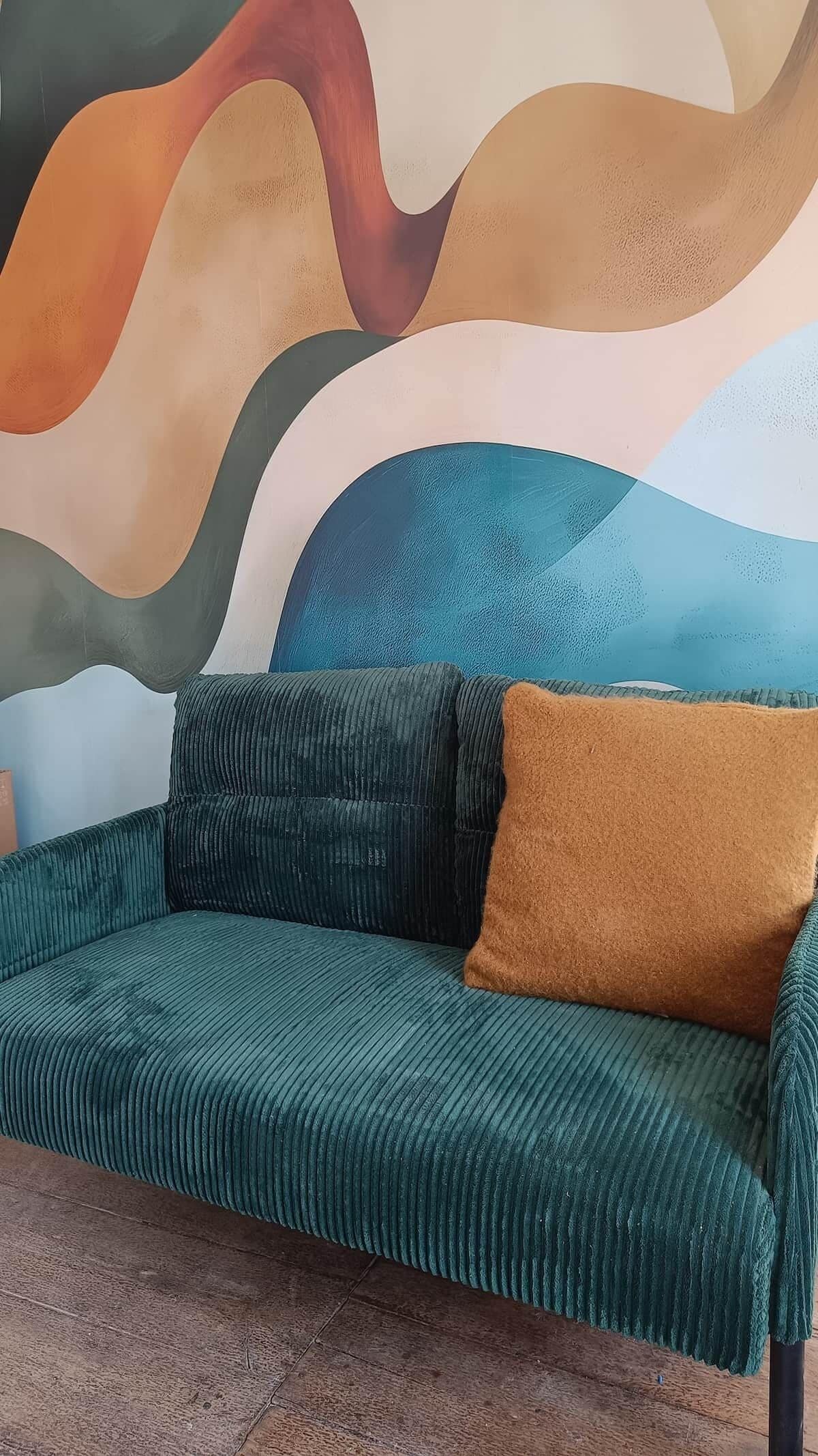 A green sofa with a brown cushion against a wall with abstract art.