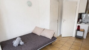 A single bed with a grey mattress and pillows, a white door, and a small kitchenette area.