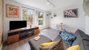 Ein modernes Wohnzimmer mit Flachbildfernseher, Holz-Multimöbel, grauem Sofa und einem Essbereich mit kleinem Tisch und Stühlen.
