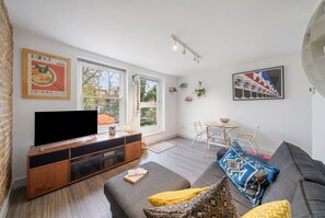 Un salon moderne doté d’un téléviseur à écran plat, d’un meuble de rangement en bois, d’un canapé gris et d’un coin repas avec une petite table et des chaises.