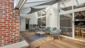 Um alpendre coberto com parede de tijolos, piso de madeira, um sofá cinza, uma cadeira de vime e uma mesa de jantar com cadeiras.