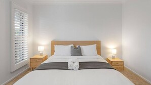 Cama bem arrumada com cabeceira de madeira, duas mesas de cabeceira com abajures e uma janela com persianas.