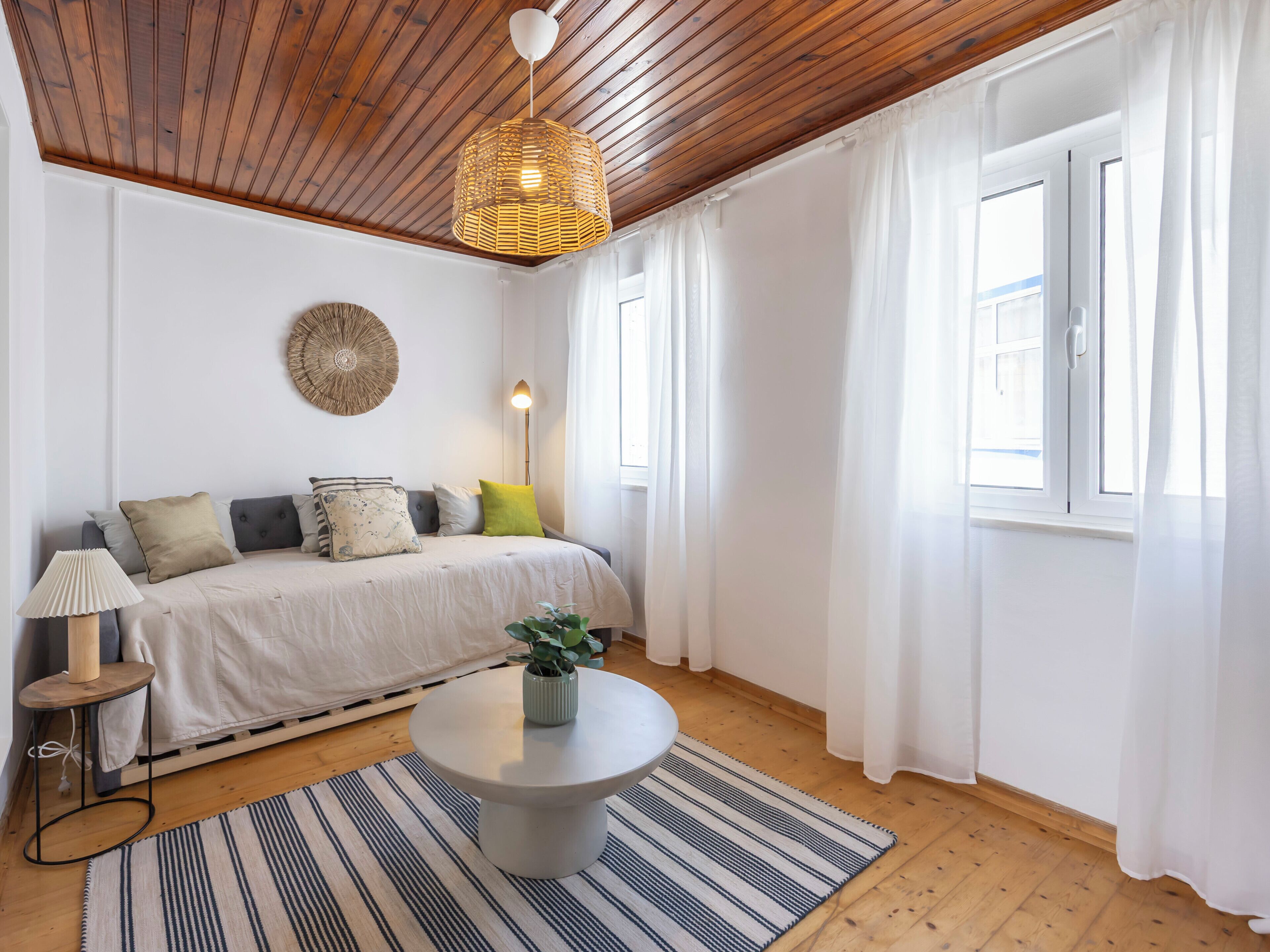 A room with a wooden ceiling, a bed with multiple pillows, a bedside lamp, a round table with a plant, and a striped rug.