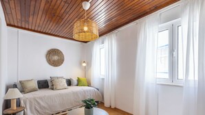 A room with a wooden ceiling, a bed with multiple pillows, a bedside lamp, a round table with a plant, and a striped rug.