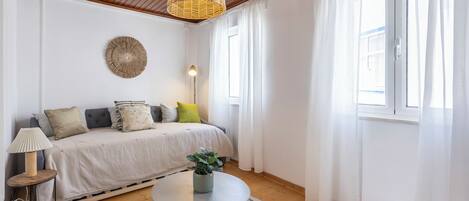 A room with a wooden ceiling, a bed with multiple pillows, a bedside lamp, a round table with a plant, and a striped rug.