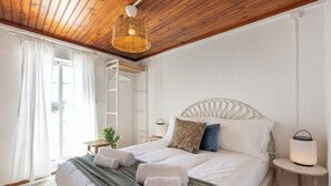 A bedroom with a wooden ceiling, a bed with white bedding, two bedside tables with lamps, and a window with curtains.