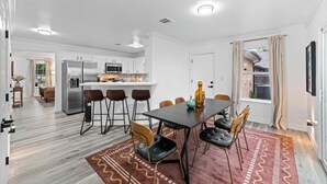 Une cuisine moderne avec des meubles blancs, un îlot central et un coin repas comprenant une table et des chaises aux couleurs sombres.