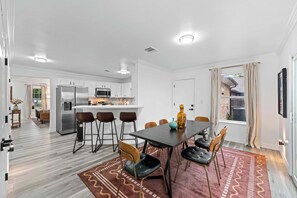A modern kitchen with white cabinets, a central island, and a dining area with a dark table and chairs. - Furnished Mid-Century 3/2 | Pool · Cowboys Stadium (Arlington)