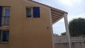 Un edificio amarillo con dos ventanas, una con contraventanas azules y una estructura de balcón blanca.