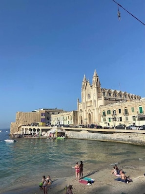 Un edificio costiero con una torre imponente e degli archi, accanto a una spiaggia con persone e una barca verde.