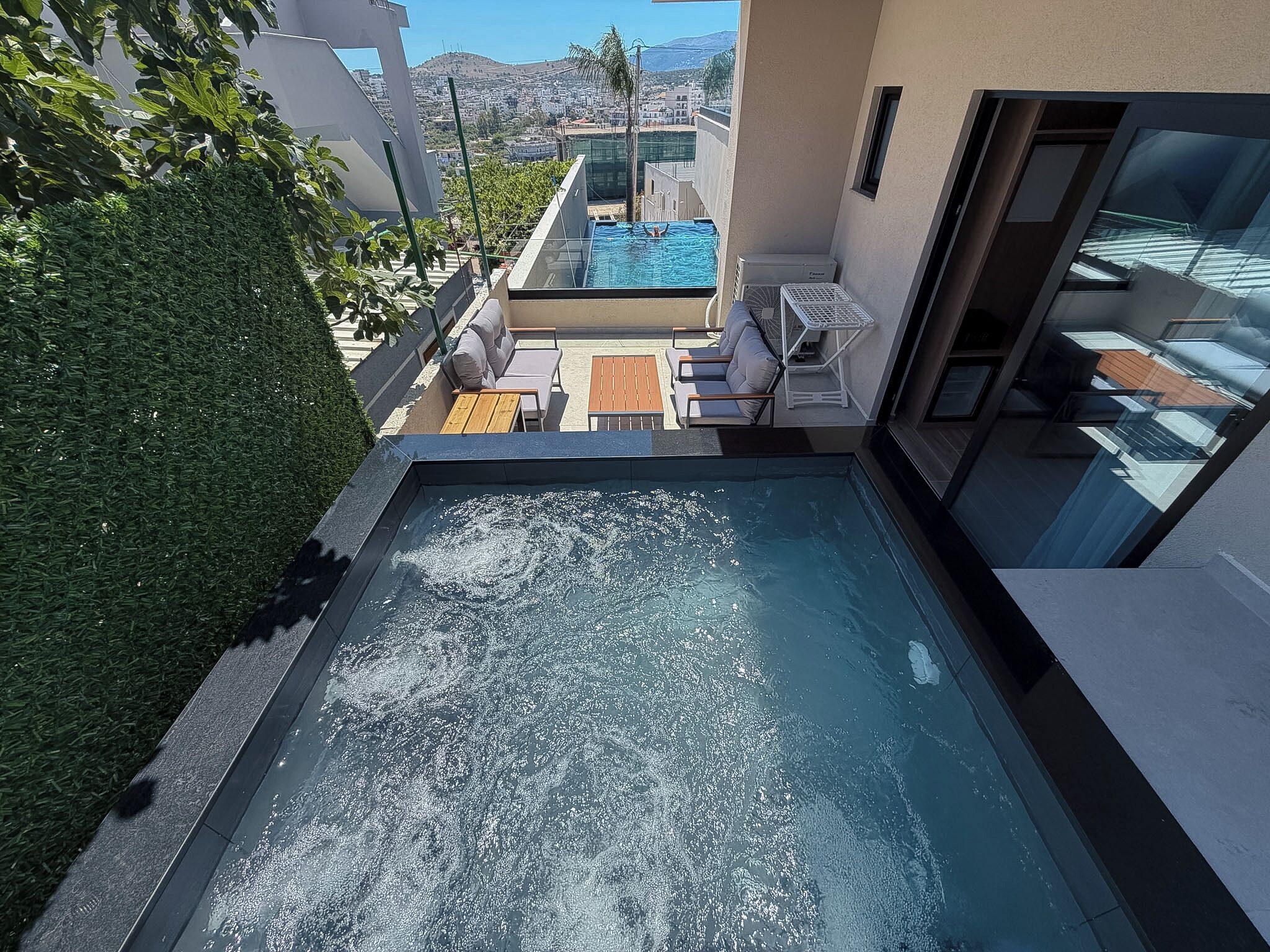 Piscina en la azotea con vistas a un paisaje urbano, rodeada de follaje y mobiliario moderno para exteriores.