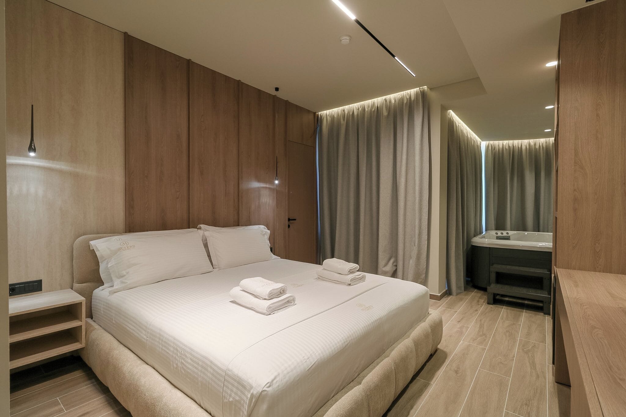 Une chambre d’hôtel moderne avec un grand lit, une table de chevet, une banquette intégrée, une petite salle de bain avec un miroir et un rideau.