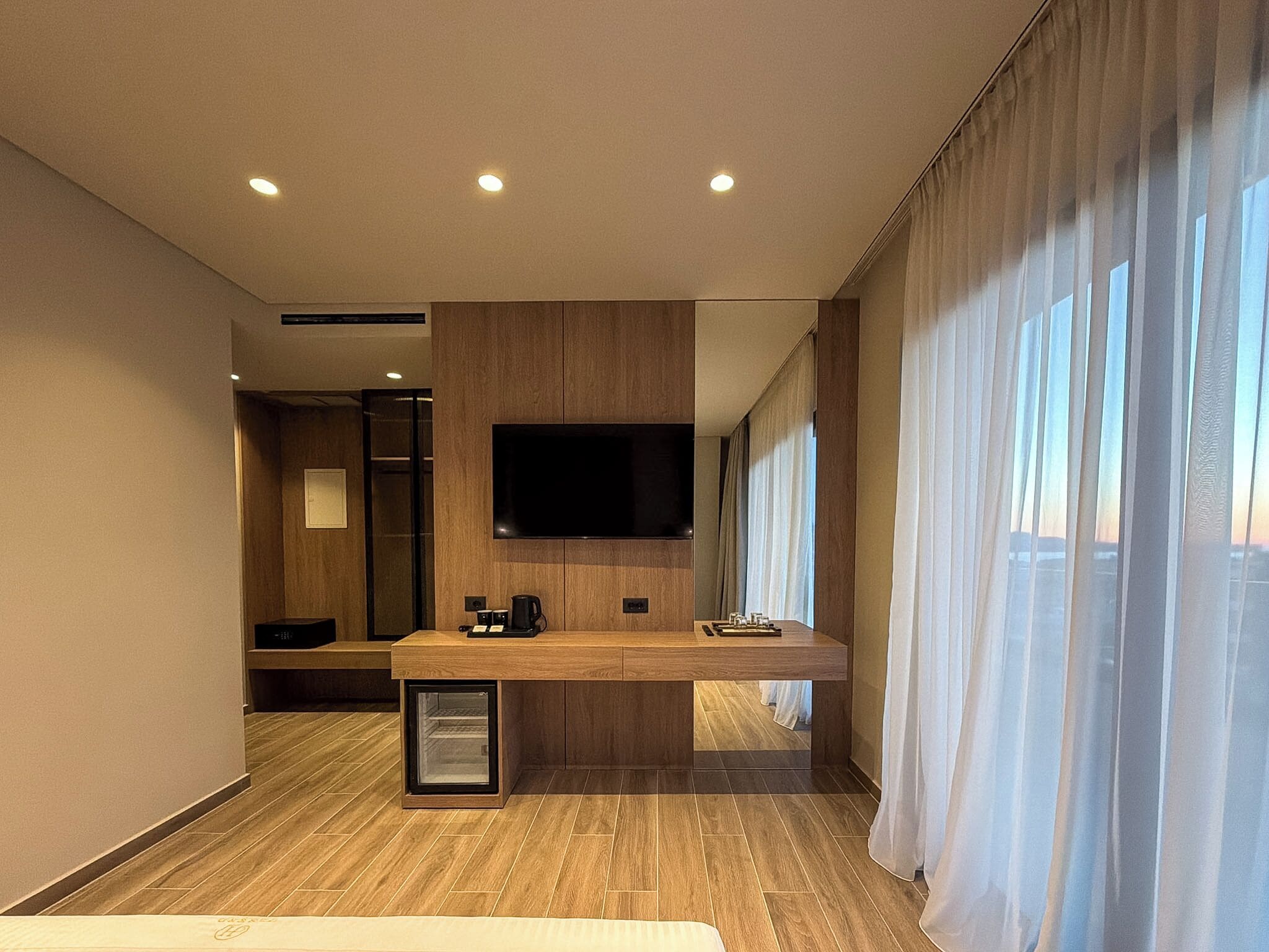 Habitación de hotel moderna con suelo de madera, televisor de pantalla plana en la pared, una pequeña estación de café y un ventanal con cortinas.