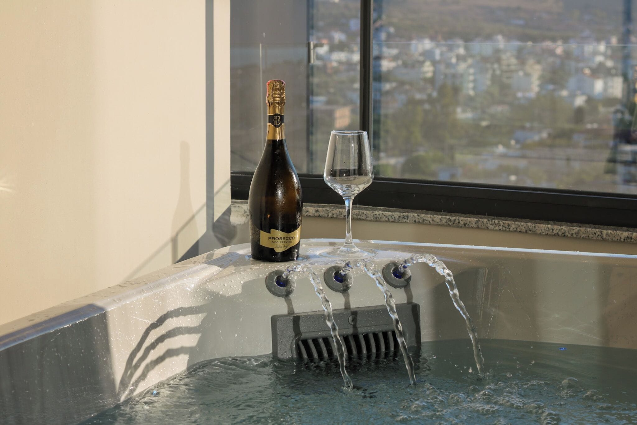 Una botella de vino Prosecco DOC Treviso junto a una copa en un alféizar con vistas a un paisaje urbano.