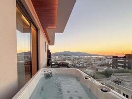 A balcony with a hot tub, a table, and chairs, overlooking a cityscape at sunset.
