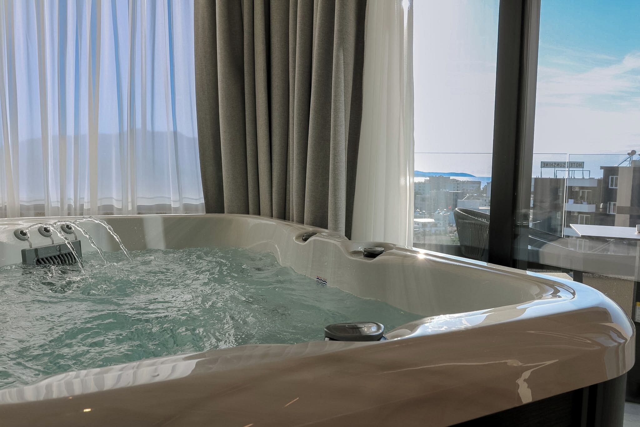 A hot tub with water jets, a window with a view of buildings, and a curtain.