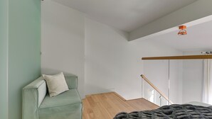 Ein schlicht eingerichteter Raum mit einem grünen Sofa, Holzboden und einem Bett mit dunkler Decke.