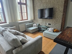Ein Wohnzimmer mit einer Couch, einem Couchtisch, einem an einer strukturierten Wand montierten Fernseher und einem Fenster mit Vorhängen.