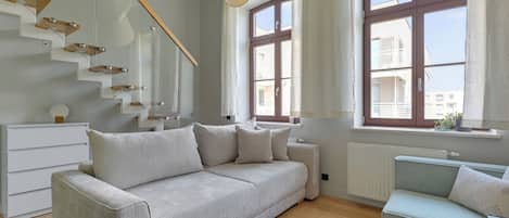 A modern living room with a sofa, a smaller armchair, a staircase, and large windows.
