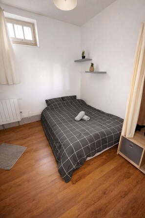 A bedroom with a bed, a radiator, a window, and a small shelf with plants.