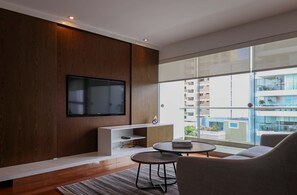 A modern living room with a flat-screen TV, a sofa, a coffee table, and a view of a building through large windows. - w Deluxe 3BR Duplex w Balcony in Miraflores (Lima)