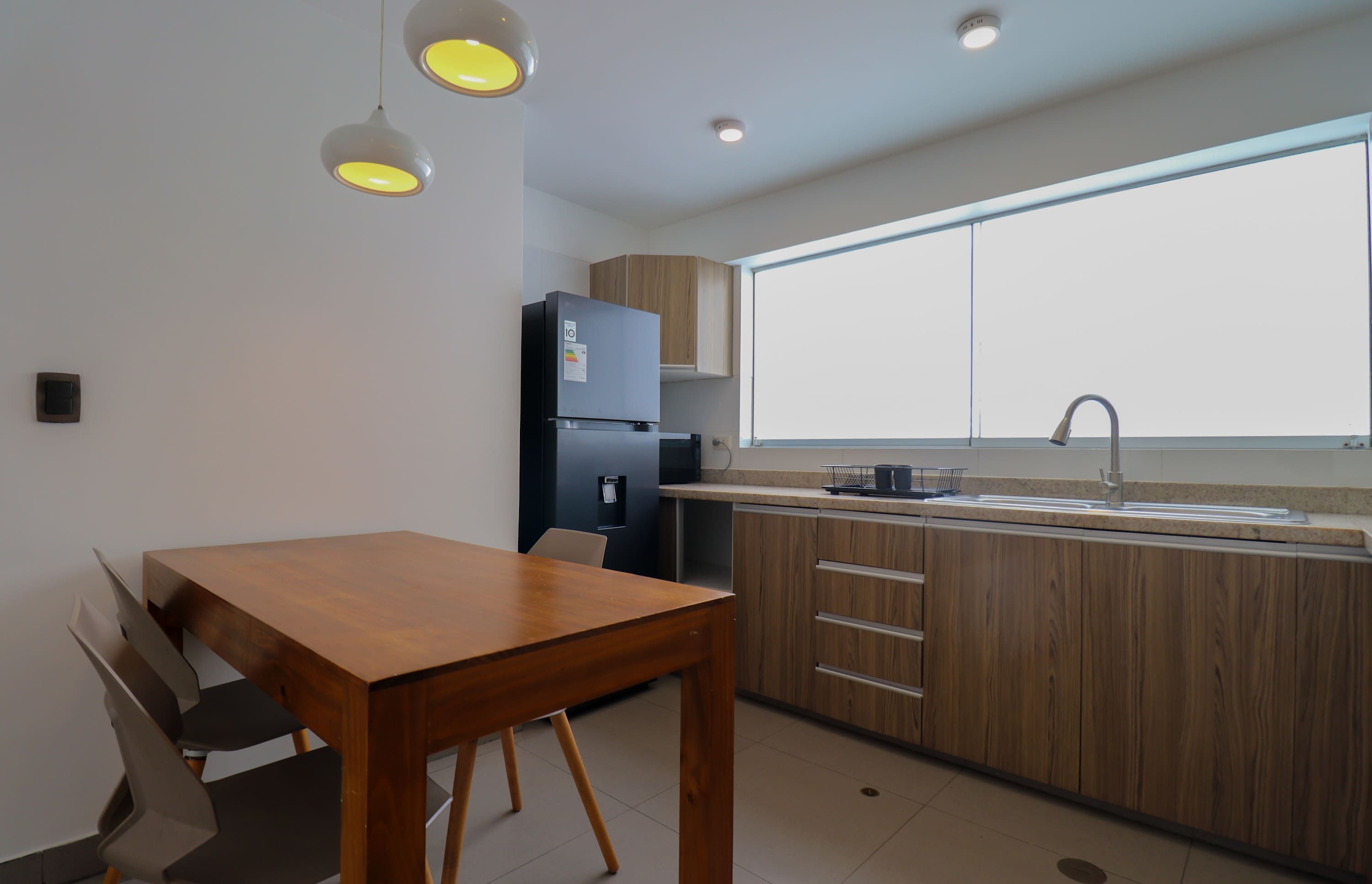 Una cocina moderna con una mesa de madera, armarios empotrados, un refrigerador y un ventanal que proporciona luz natural.