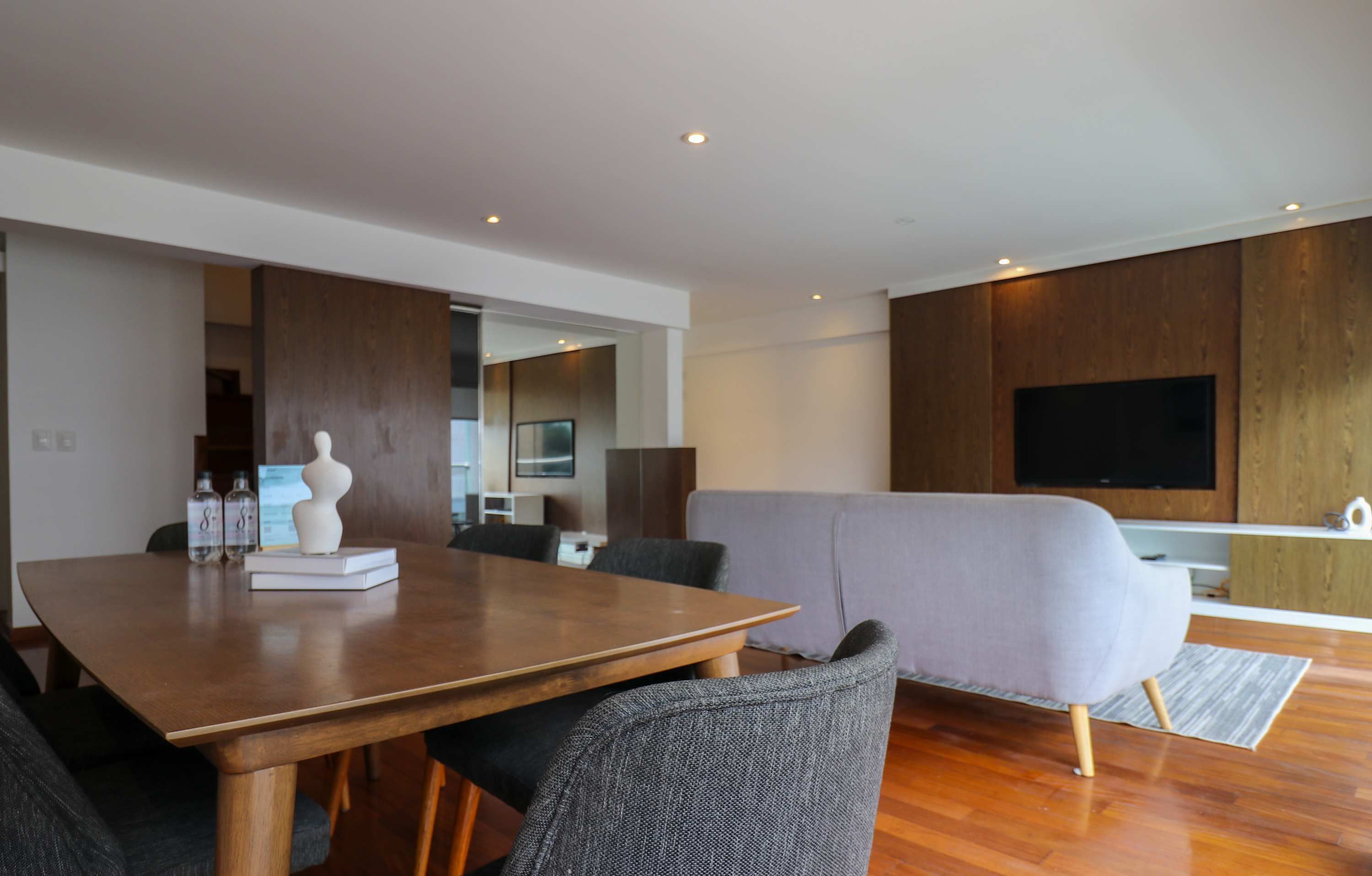 A modern living room with a wooden dining table, grey sofa, and a flat-screen TV mounted on the wall.