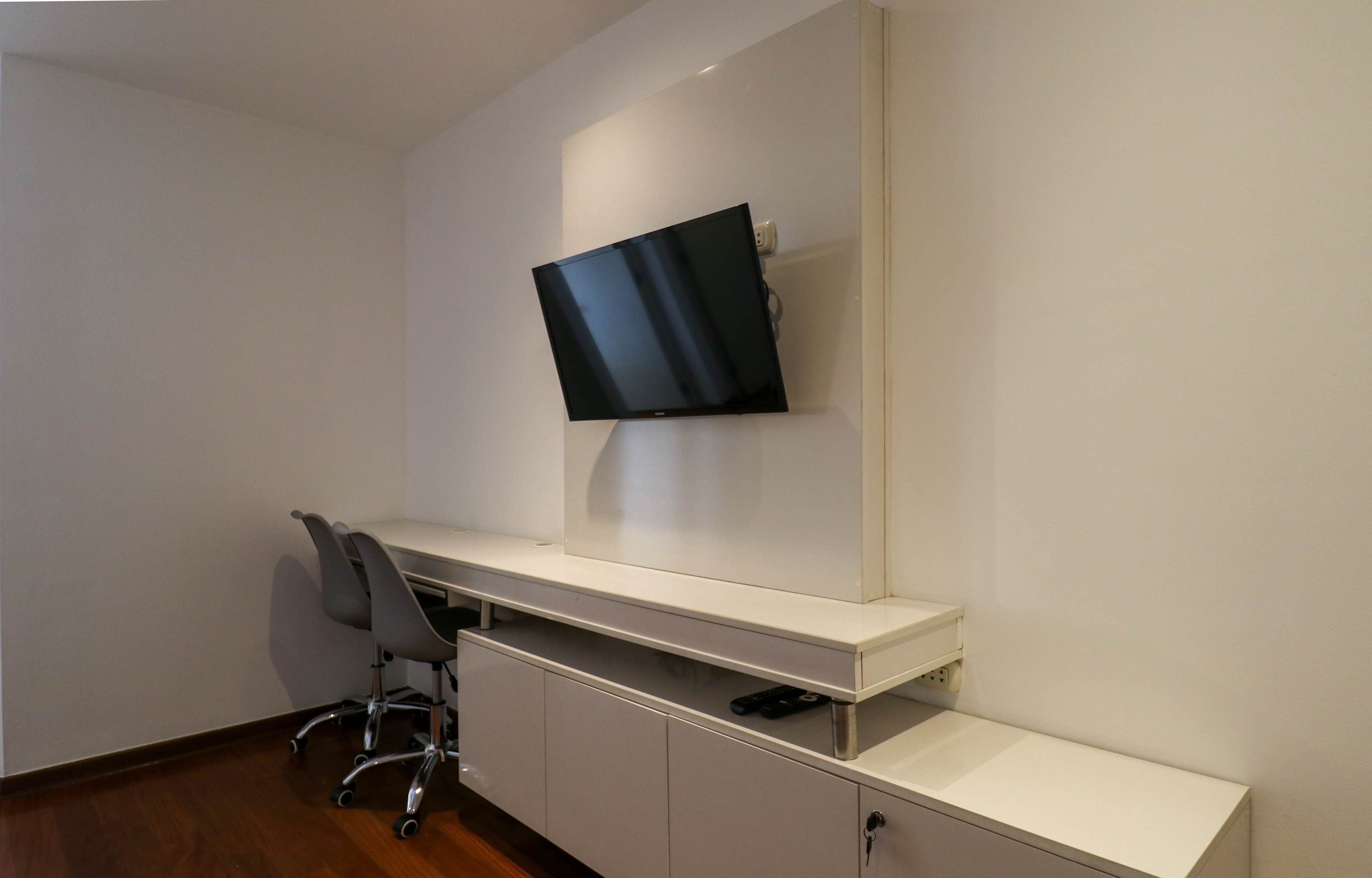 A room with a white desk, a chair, a mounted TV, and a cabinet.