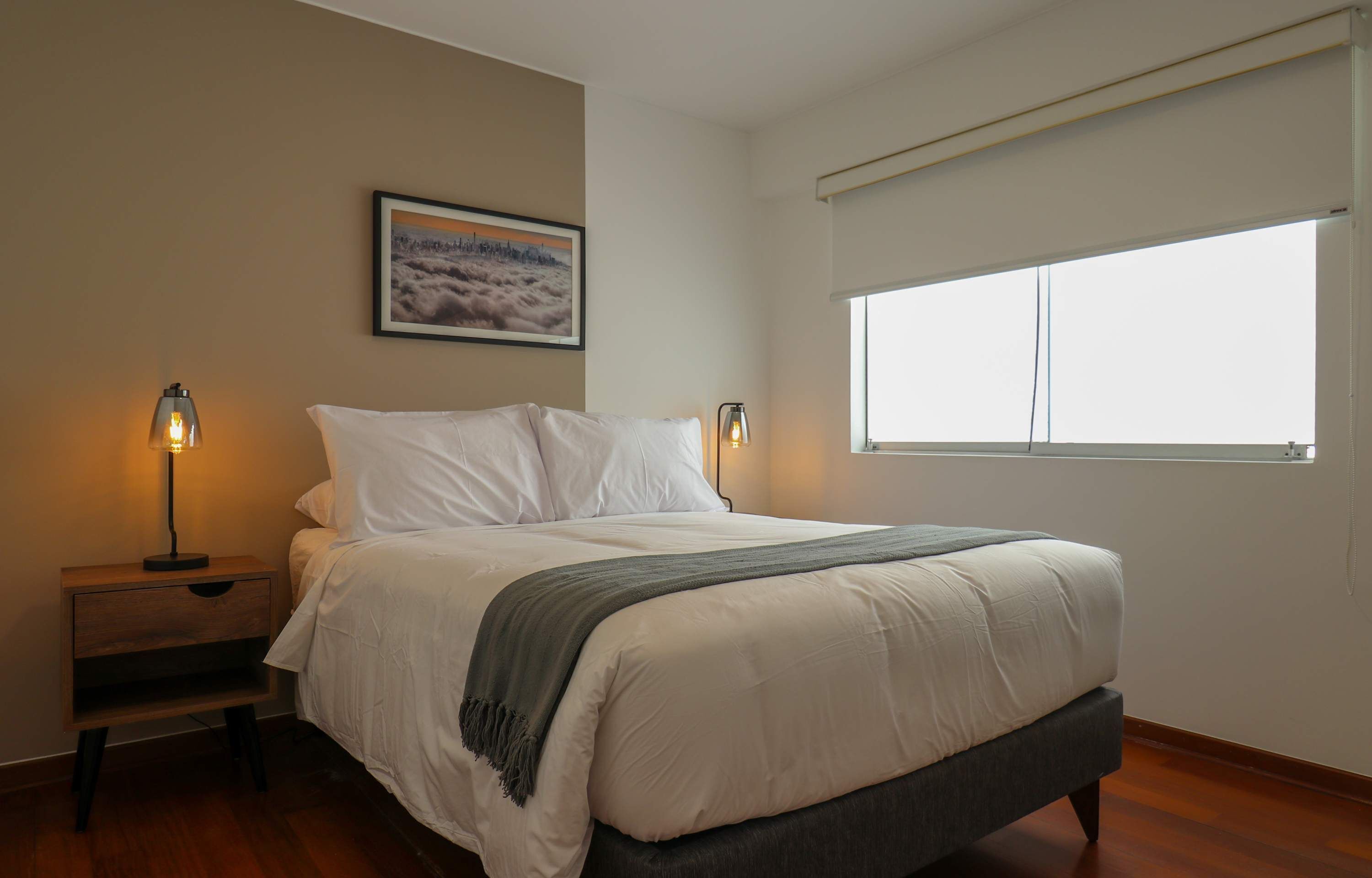 A bedroom with a bed, two bedside lamps, a nightstand, a window, and a framed picture on the wall.
