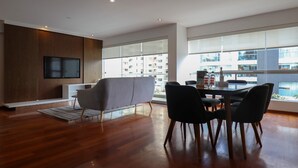 Een moderne woonkamer met een houten vloer, een grijze bank, een eettafel met stoelen, een aan een houten wand bevestigde televisie en grote ramen met uitzicht op een gebouw.