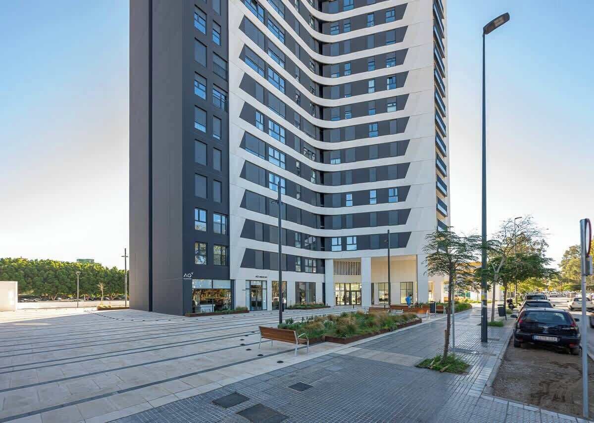 Un edificio moderno con fachada curva, una plaza pavimentada con bancos y una calle con coches aparcados.