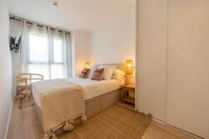 A bedroom with a bed, bedside lamps, a wicker chair, and a window with curtains. - Zapotlan - City View Malaga Apartment w/ Pool & Gym (Málaga)