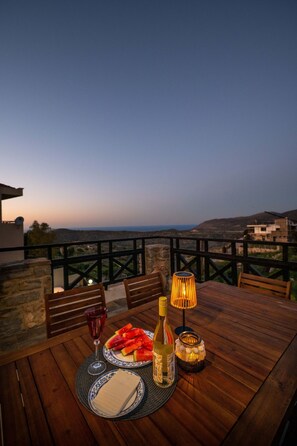 A wooden table with a bottle, a glass, and a plate of food on a balcony overlooking a landscape. - Altair Blue Villa - Private Jacuzzi and Views (Hersonissos)