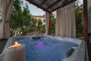 A hot tub with a candle, surrounded by a pergola and curtains, with a view of a garden and buildings in the background. - Altair Blue Villa - Private Jacuzzi and Views (Hersonissos)