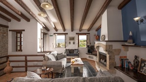 A cozy living room with exposed wooden beams, a stone fireplace, and a view of the outdoors. - Altair Blue Villa - Private Jacuzzi and Views (Hersonissos)