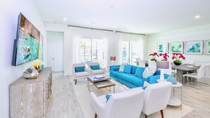 Ein modernes Wohnzimmer mit einem blauen Sofa, weißen Sesseln, einem Holzschrank und einem Essbereich mit Tisch und Stühlen.