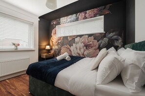 Un dormitorio con una cama grande, un mural floral en la pared, una ventana con persianas, una mesita de noche con lámpara y un radiador.